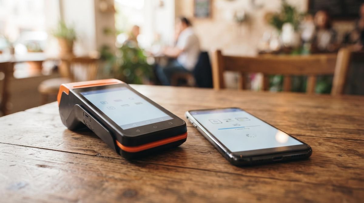 Uma maquininha de cartão moderna posicionada sobre uma superfície de madeira ao lado de um smartphone com o aplicativo de pagamentos aberto