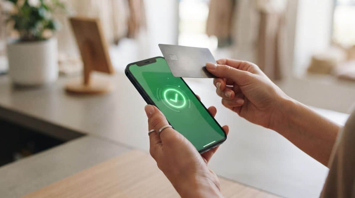 Close-up das mãos de uma pessoa segurando um iPhone com a tela exibindo confirmação de pagamento aprovado, com um cartão de crédito sendo aproximado